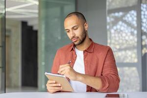 A young, focused entrepreneur is writing notes in a notebook at his desk, showcasing concentration and diligence in the office. photo