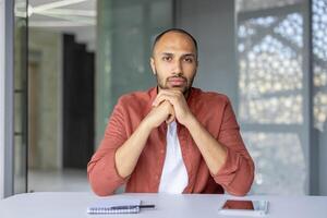 A serious man looks directly at the camera while sitting at a desk, appearing to be on a call photo
