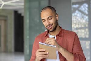 A young man is seen writing in a notepad with a pen, looking focused and content, probably at work. photo