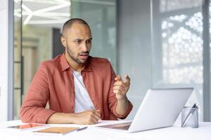 Serious and focused man talking to colleagues and partners sitting at desk, using laptop for call. Businessman inside office at workplace. photo