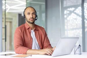 A focused call center agent, wearing a headset, looks at the camera while sitting at a desk. The man works on a laptop in an office environment. photo