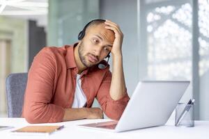 Sad bored man with headset working inside office with laptop, company employee dissatisfied and unhappy at work, consulting clients online. photo
