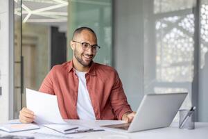 A smiling man reviews documents while working on his laptop in a modern office setting, suggesting remote work or online collaboration. photo