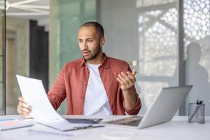 A surprised man examines documents while sitting at a desk. A laptop and office supplies are present, too. He seems bewildered by the information. photo