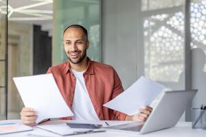 Successful businessman doing paperwork, man in casual clothes checking documents and bills, satisfied with results of activity smiling, working inside office with laptop. photo
