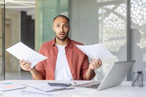 A surprised man holds paperwork while working at his desk, possibly experiencing financial stress. photo