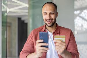 Close-up photo, man with phone and bank credit card smiling makes online purchases in online store remotely. Businessman inside office at workplace transfers money. photo