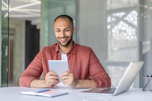 Successful male businessman using tablet computer sitting at desk inside office. Company employee satisfied with achievement smiling, using online financial app. photo