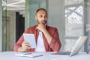 A thoughtful man at a desk holds a tablet, pondering ideas while working on a laptop in a modern office setting. photo