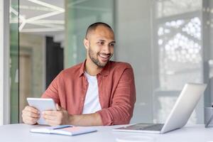 A smiling man sits at a desk, using a laptop and holding a tablet. Natural light floods the modern workspace, enhancing the scene photo