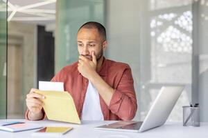 A concerned man is looking at a letter or document, his expression suggesting worry or concern. He is in an office setting with a laptop. photo