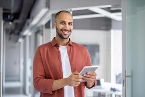 A smiling, bald man in a red shirt holds a tablet in an office setting with a blurred background. photo