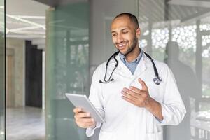 Young doctor consults patients remotely using tablet computer and call. Man in white medical coat talking while standing inside office of modern clinic. photo