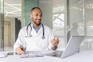A doctor wearing a lab coat smiles while talking during a conference from his office. He's using a laptop and gesturing. photo