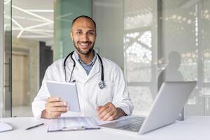 Portrait doctor inside the office at his workplace inside the office. A man in a white medical coat smiles and looks at the camera, holds a tablet computer in his hands, consults patients remotely. photo