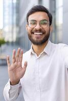A man with a beard and glasses waves during a call outside a building photo