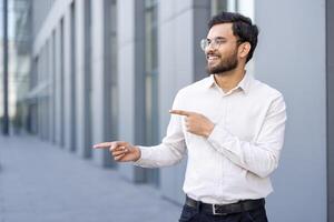 A smiling businessman of Indian descent points sideways, wearing glasses and a button-down shirt, outdoors with a modern building in the background. photo