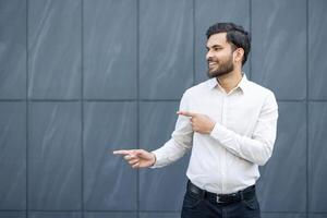 A smiling man in a white shirt points to the side against a backdrop of a gray tiled wall. photo