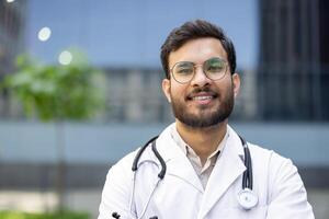 A smiling Indian doctor in a white coat and glasses stands outside. He has a stethoscope around his neck, with a building visible in the background. photo