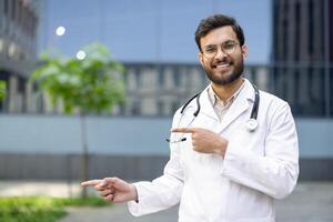 A smiling male doctor in a white coat points to the side with a stethoscope around his neck. He is in front of an office building. photo