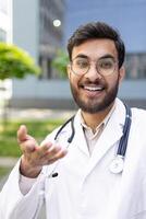 A smiling doctor with a stethoscope gestures towards the viewer while talking on a call outside, offering a welcoming hand gesture photo