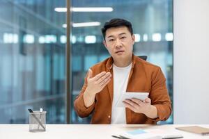 An Asian man in a business setting gestures while holding a tablet, seemingly explaining something. photo