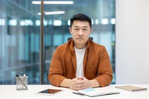 A focused Asian man sits at a desk, looking directly at the camera in a modern office setting, wearing a button-down shirt. photo
