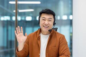 A cheerful Asian man wearing a headset waves at the camera, smiling warmly, in a bright office setting. photo