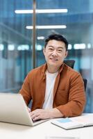 An Asian man smiling while working on a laptop in a modern office environment. photo