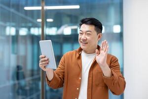 An Asian man waves while holding a tablet, likely during a call. He's smiling warmly in an office setting. photo