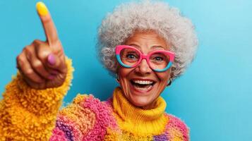 Joyful older woman with colorful sweater and glasses pointing against a bright blue background in a cheerful expression celebrating life and positivity photo