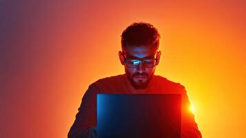 Man working on laptop in dimly lit room with vibrant orange and blue background at night, focused on task while wearing glasses photo
