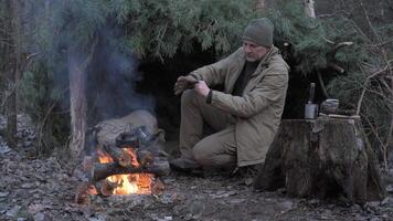 Man preparing tea by campfire near pine shelter in cold forest, A camper tends to a kettle on a fire as he rests beside his rustic shelter in the wilderness video