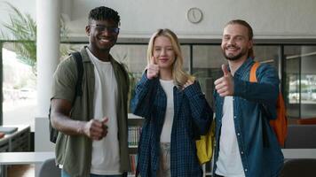 Closeup three multiracial friends diversity students smiling showing thumbs up likes recommend success achievement looking at camera in university corridor hall together smile positive show agree sign video