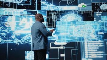 Black male IT engineer examining digital AI brain interface on dashboard, symbolizing neural networks, science and algorithmic thinking. Cybernetics for high tech machine learning. Camera A. video