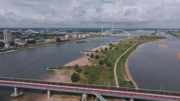 Aerial view of Nijmegen, Netherlands, showing the Waal River, De Oversteek bridge, moving water, vehicles, wind turbines, and urban landscapes. video