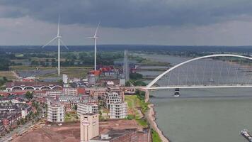 Aerial view of De Oversteek bridge in Nijmegen, Netherlands, with boats on the Waal River, green landscapes, sandy banks, and power lines. video