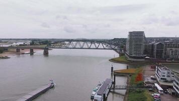 Aerial view of Nijmegen, Netherlands, featuring the Waal River, a steel bridge, a moving barge, modern buildings, and historic architecture under overcast skies. video