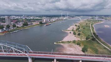 Aerial view of Nijmegen, Netherlands, showing the Waal River, De Oversteek bridge, a moving barge, wind turbines, urban areas, and green spaces. video