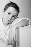 Black and white photo of a boy with curly hair, sitting on a wooden chair, hands folded in his lap. The expression is serious and contemplative, lit with classic Rembrandt lighting.
