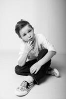 Black and white portrait of a young boy lying on his stomach, chin resting on hands, looking at the camera with a dreamy, thoughtful expression. Soft shadows create a calm mood. photo
