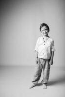 Black and white studio portrait of a small boy sitting on a stool, looking directly at the camera with a soft, innocent expression. Plain dark background, gentle lighting emphasizes facial features. photo