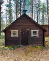 Dark wooden cabin in northern Sweden stands quietly among tall pines under a partly cloudy sky photo