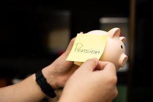 Hands attaching a yellow sticky note with the word Pension written on it to a piggy bank. Planning for Pension and Retirement Savings. saving for the future, and securing retirement. photo