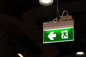 Illuminated Green Emergency Exit Sign with Arrow Pointing Left. green emergency exit sign hangs from the ceiling in a dimly lit indoor space. concepts of safety, evacuation, emergency preparedness. photo