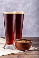 Two tall glasses of dark beer with creamy foam next to rustic clay bowl of salted peanuts. Wooden table and linen cloth accentuate cozy, traditional feel. Rustic dark beer and peanuts photo
