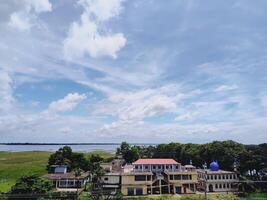 Semi urban landscape with a mosque, roofed building, lush greenery, and calm water under a partly cloudy sky. Ideal for rural development, nature, and scenic architecture themes. photo