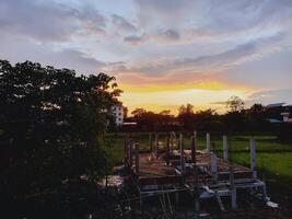 Rural construction site with visible concrete pillars and foundation set against lush green fields scattered trees and a warm sunset sky. A large tree stands silhouetted on the left. photo