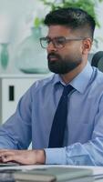 Vertical view Indian businessman Arabian business man ethnic male worker guy professional entrepreneur working in office typing computer checking documents work focused job online at workplace table video