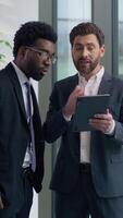 Vertical view two diversity multiethnic business men colleagues discuss online success looking in digital tablet amazed good news multiracial businessmen males talking in office about startup growth video